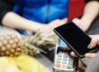 A customer uses a smartphone for contactless payment at a retail checkout with fresh produce.