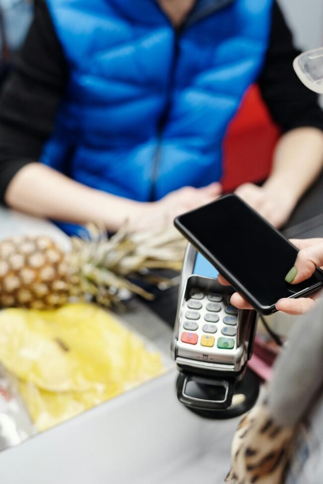Photo by Jack Sparrow A customer uses a smartphone for contactless payment at a retail checkout with fresh produce.