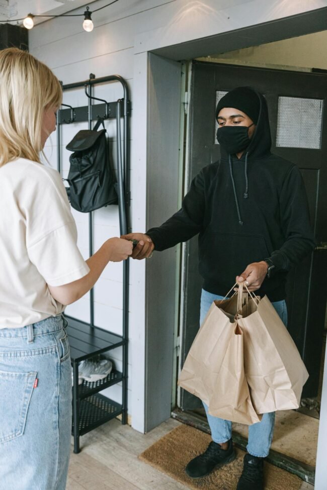Photo by Ivan Samkov A masked courier hands over shopping bags to a customer in a contactless delivery exchange.